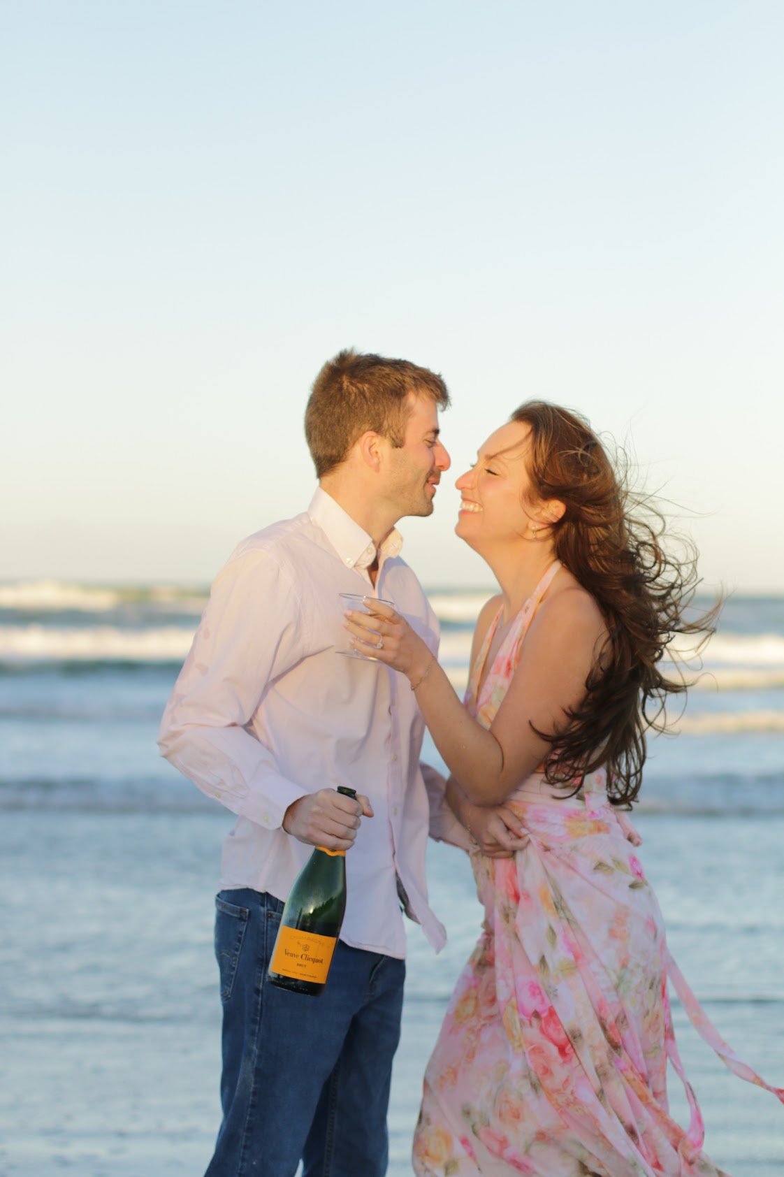 Engaged couple laughing with a bottle of champagne on the beach at sunset